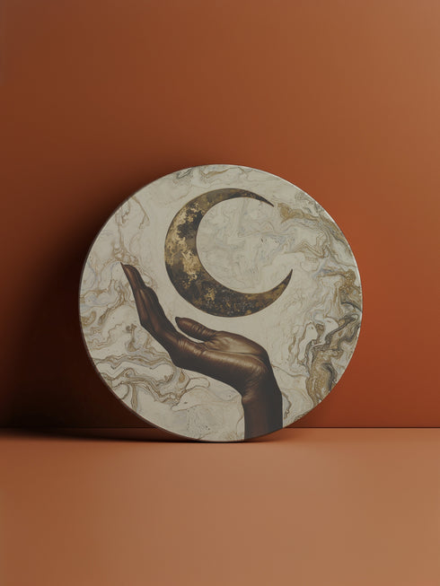 A ceramic coaster featuring a bronze-colored hand reaching toward an antiqued gold crescent moon against a swirling ivory and gold marble background, available in round and square shapes with cork backing.